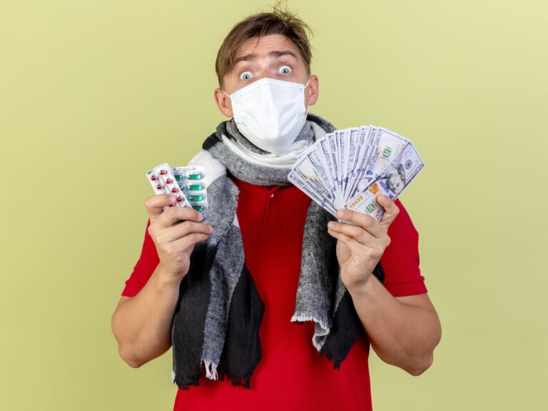 impressed young handsome blonde ill man wearing mask holding money packs medical pills isolated olive green wall wondring why are prescription drugs so expensive