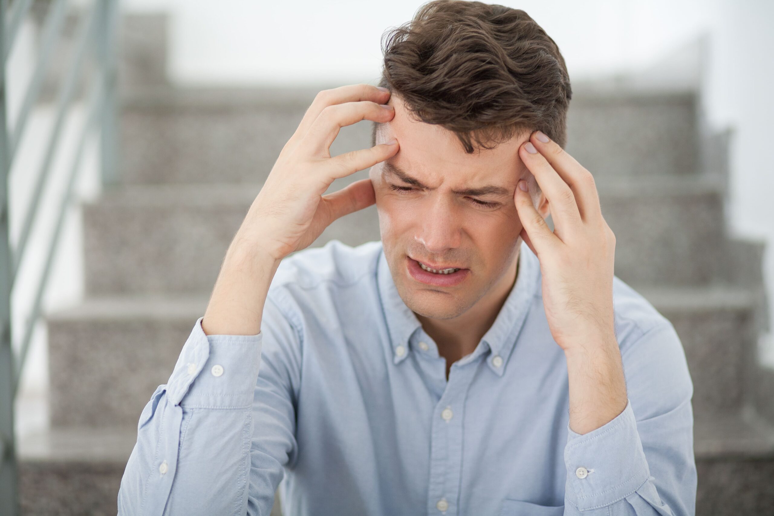 Man sitting on indoor stairs holding his temples in pain, showing symptoms of a severe headache and stress associated with REN migraine treatment.