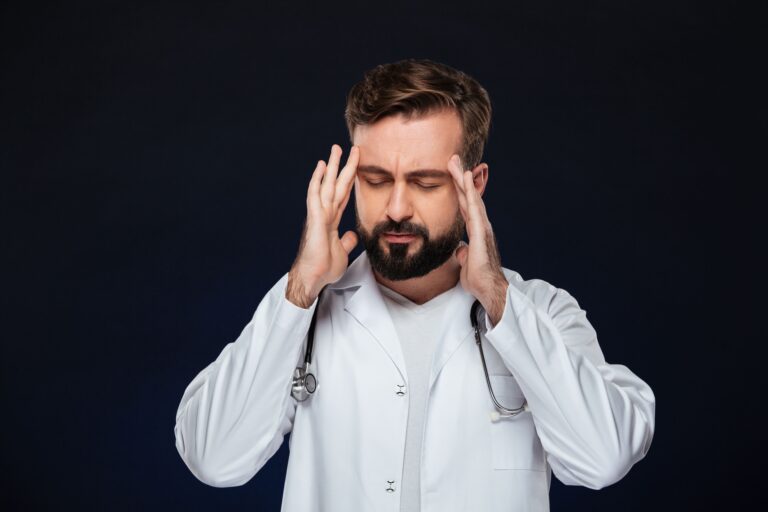 Doctor in white coat holding his temples with eyes closed against a dark background, illustrating severe facial pain and stress related to trigeminal neuralgia treatment.