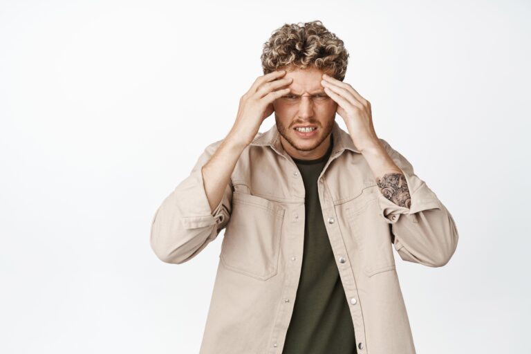 Young man grimacing and pressing hands to temples experiencing migraine postdrome symptoms and headache pain