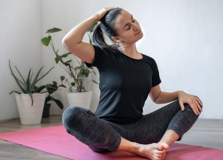 Woman practicing gentle neck stretches while seated on a yoga mat at home, demonstrating yoga for migraine prevention and relaxation exercises to reduce tension and stress.