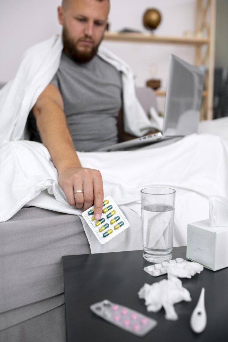 A sick man sitting in bed reaches for a blister pack of capsules beside a glass of water, tissues, and medication on a bedside table. The image illustrates chronic pain management and the potential complications of long-term opioid use, including opioid induced hyperalgesia.
