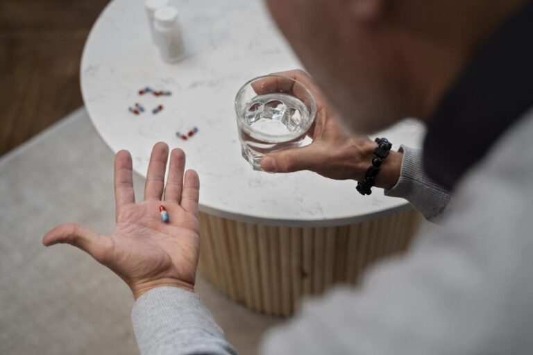 A close-up view of an adult holding a red-and-blue capsule in one hand and a glass of water in the other, seated beside a table with additional medication capsules and a prescription bottle. The image illustrates responsible medication use, patient decision-making, and opioid therapy considerations in the context of the 2022 CDC opioid prescribing guidelines, emphasizing informed treatment choices, safe prescribing practices, and patient-centered care.