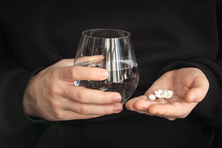Close-up of a person holding a glass of water in one hand and several white tablets in the other against a dark background, illustrating medication use during Lofexidine Opioid Withdrawal treatment and symptom management.