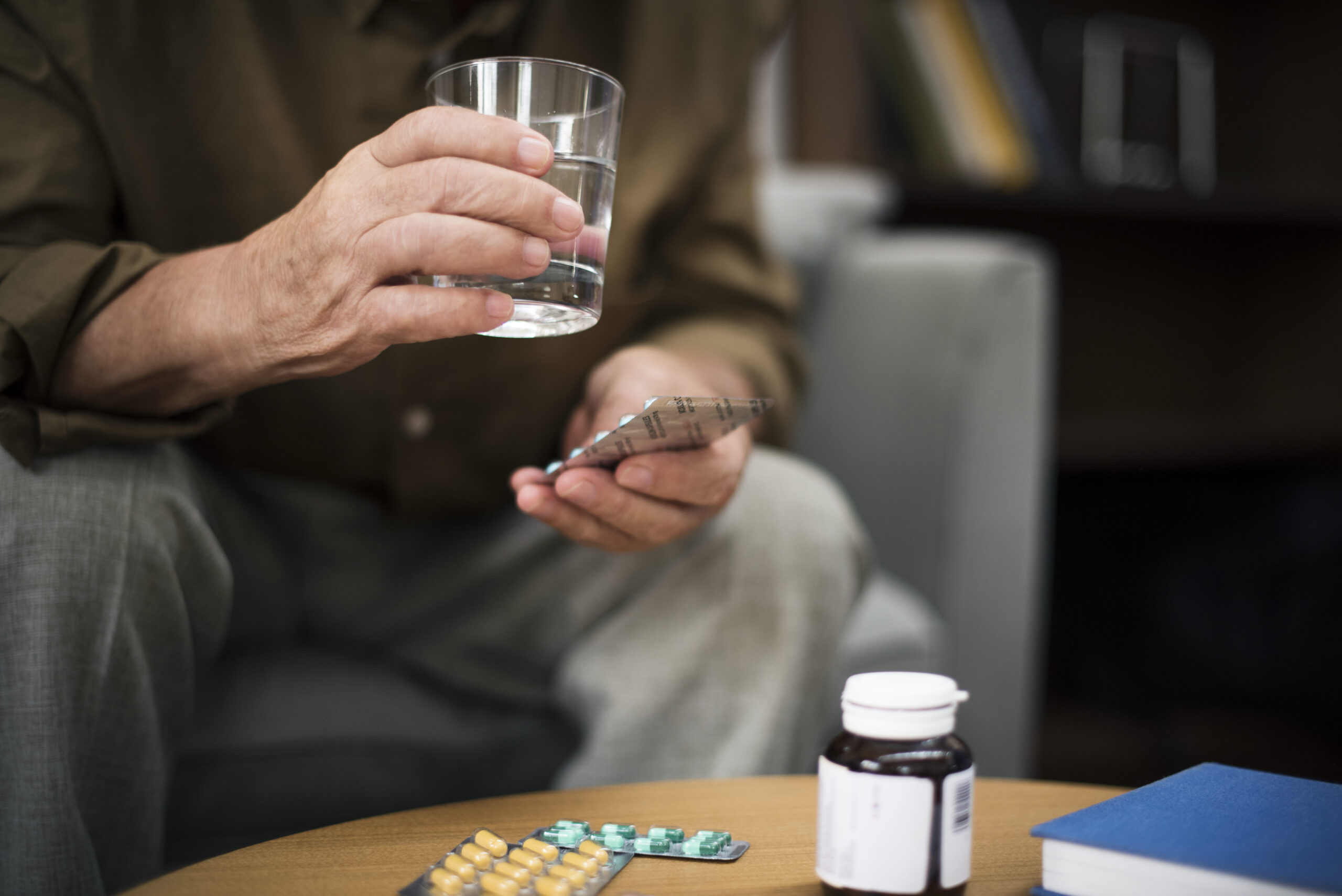 Older adult holding a glass of water and a medication blister pack while seated at home, with prescription bottles and pills on a table, illustrating Medicare opioid coverage policy and access to prescription pain medications.