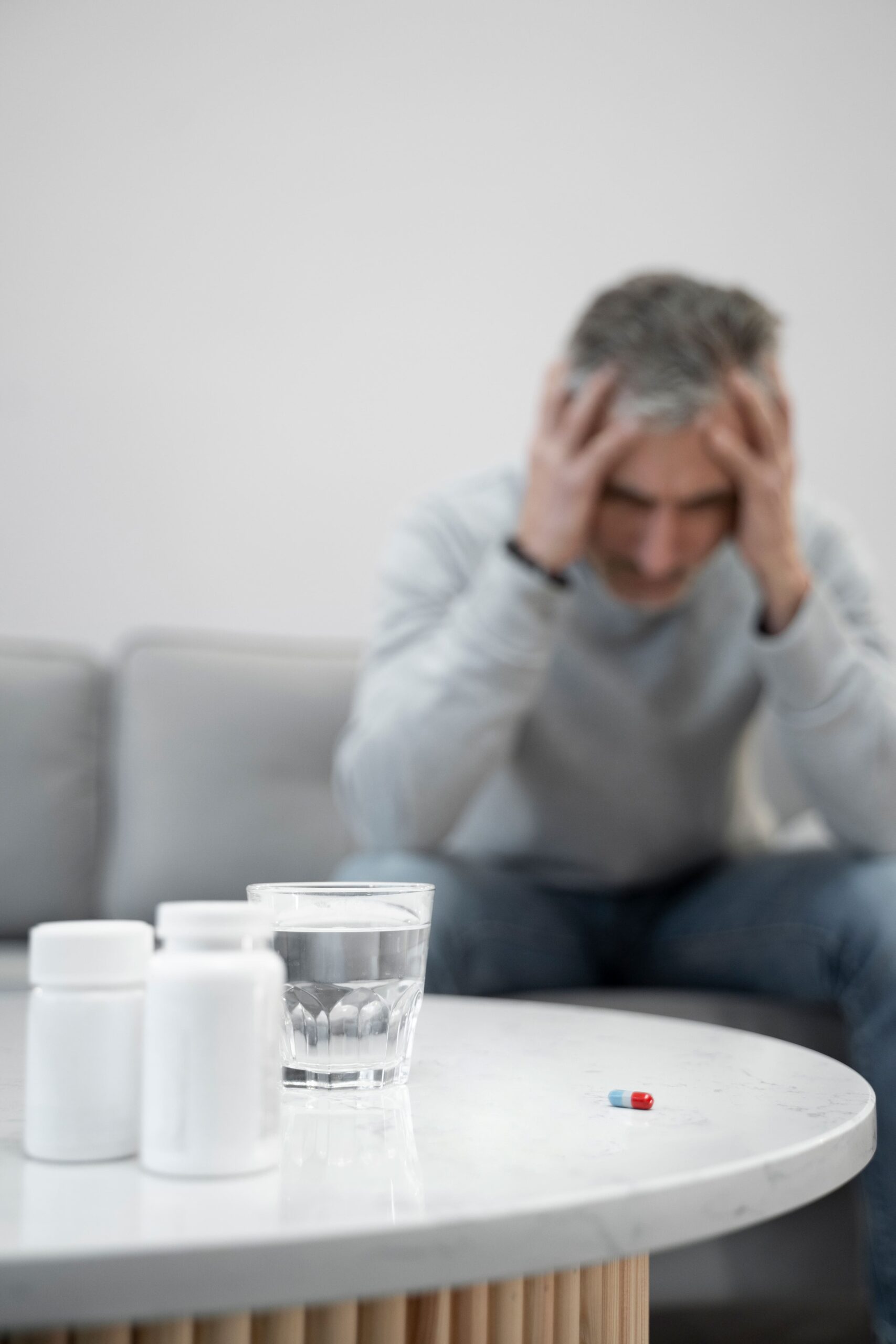 Middle-aged man sitting on a couch with his head in his hands, appearing distressed, while pill bottles, a glass of water, and a capsule rest on a table in the foreground, illustrating the challenges of opioid recovery and the role of mind body therapy opioid use in managing pain and emotional well-being.