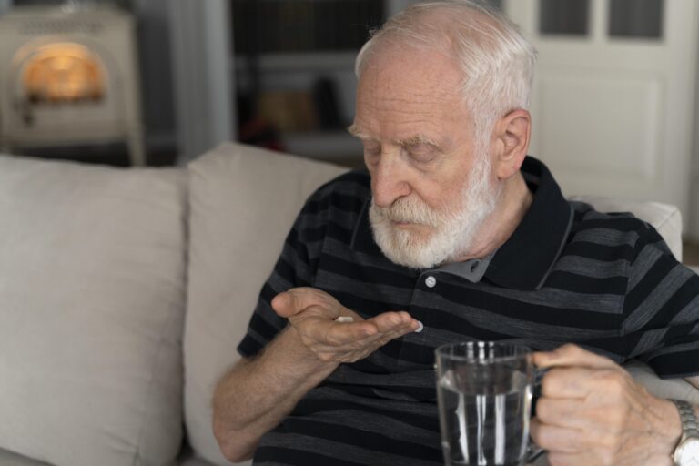 Elderly man sitting on a couch at home holding medication pills in his hand and a glass of water, illustrating concerns related to opioid use disorder in elderly patients and prescription medication management.