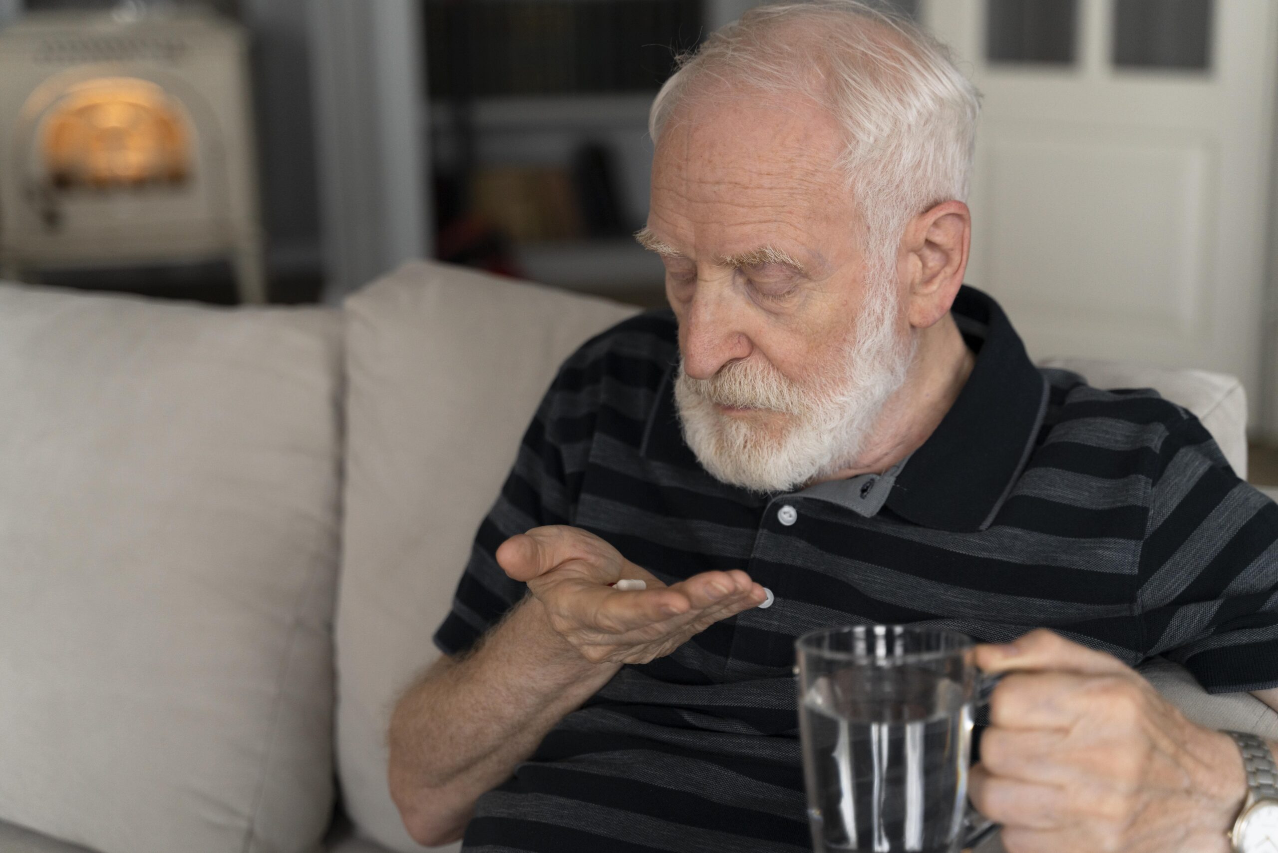 Elderly man sitting on a couch at home holding medication pills in his hand and a glass of water, illustrating concerns related to opioid use disorder in elderly patients and prescription medication management.